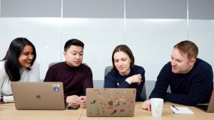Four workers seated at a table, facing us, with a woman on the left, then a man, a woman, and another man. The man on the far right has a coffee cub and a notepad and pen on the table. The group is looking at two open computers, one of which has many stickers on its back.