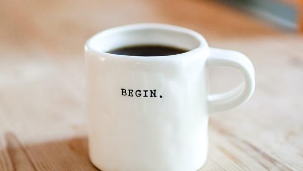 a white ceramic coffee cup, filled with coffee, with its handle to the right with a design of typewritten letters BEGIN.